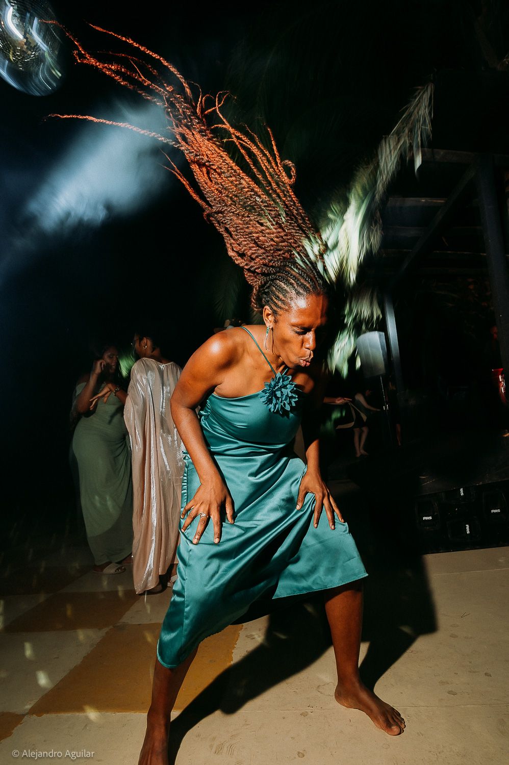 A woman in a teal satin dress dances energetically with dramatic braided hair flowing upward at a nighttime event.
