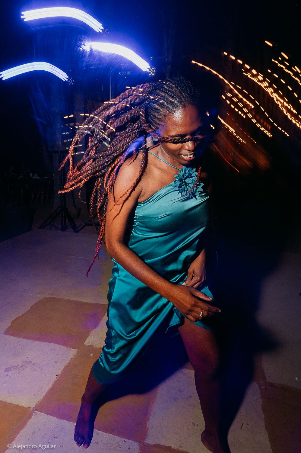 Woman in teal dress dancing joyfully with braided hair flying, illuminated by colorful light trails at a nighttime party.
