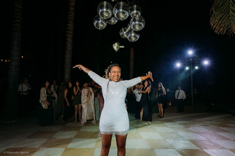 Bride in white mini dress joyfully tosses bouquet at outdoor evening wedding reception with guests watching.