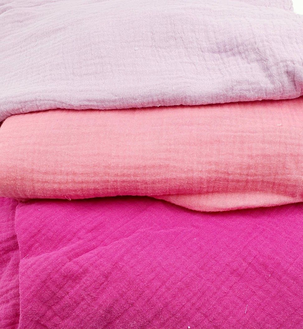 Stack of three folded muslin blankets in light pink, coral, and magenta shades.