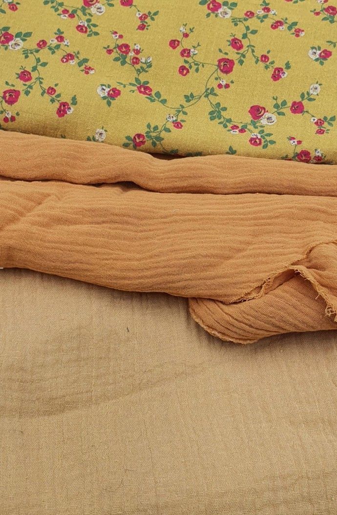 Three layered fabrics including yellow floral cotton, burnt orange muslin, and natural beige linen textile samples.