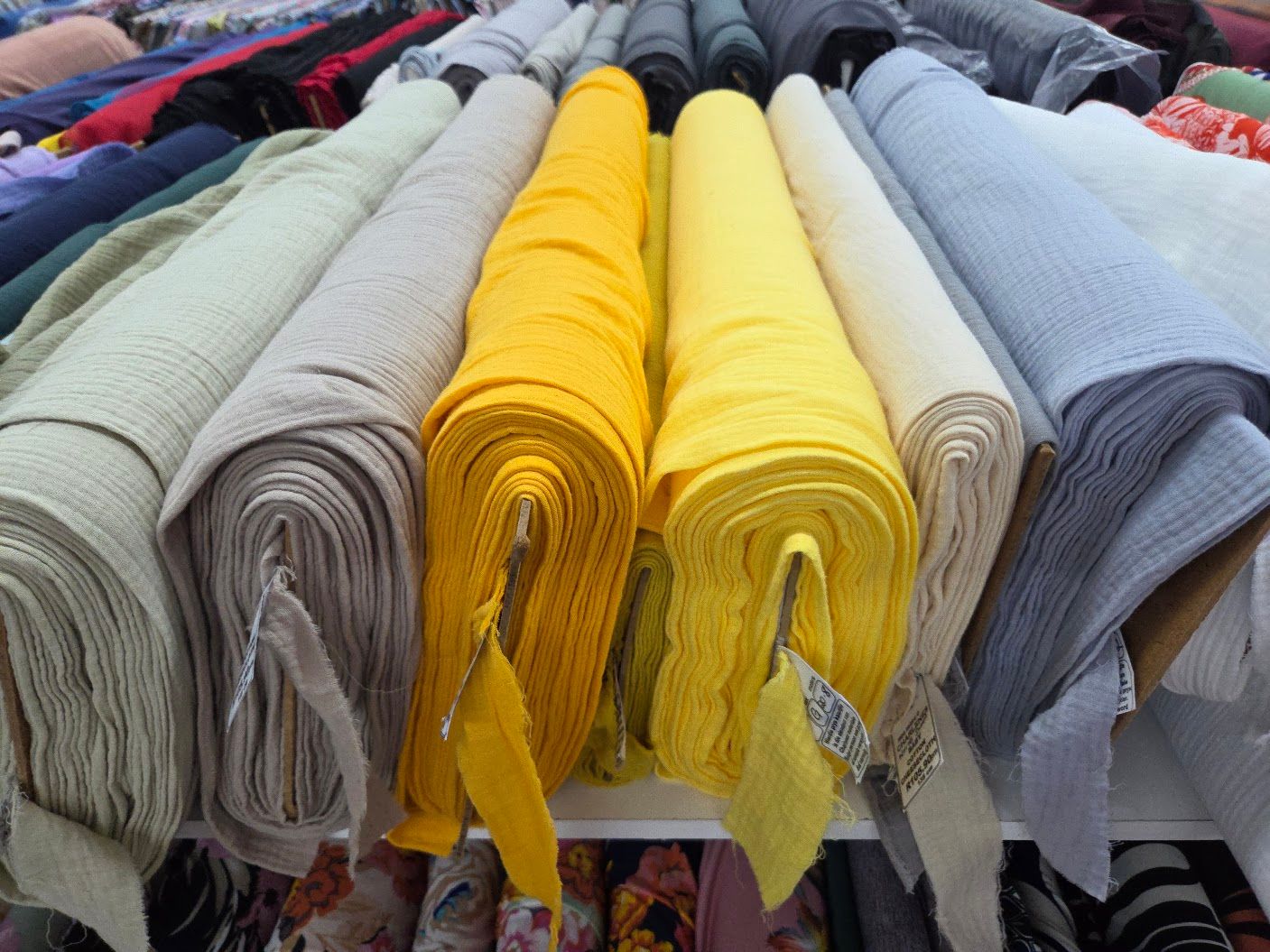 Rolls of colorful fabric bolts in yellow, gray, and beige displayed on shelves at a textile market.