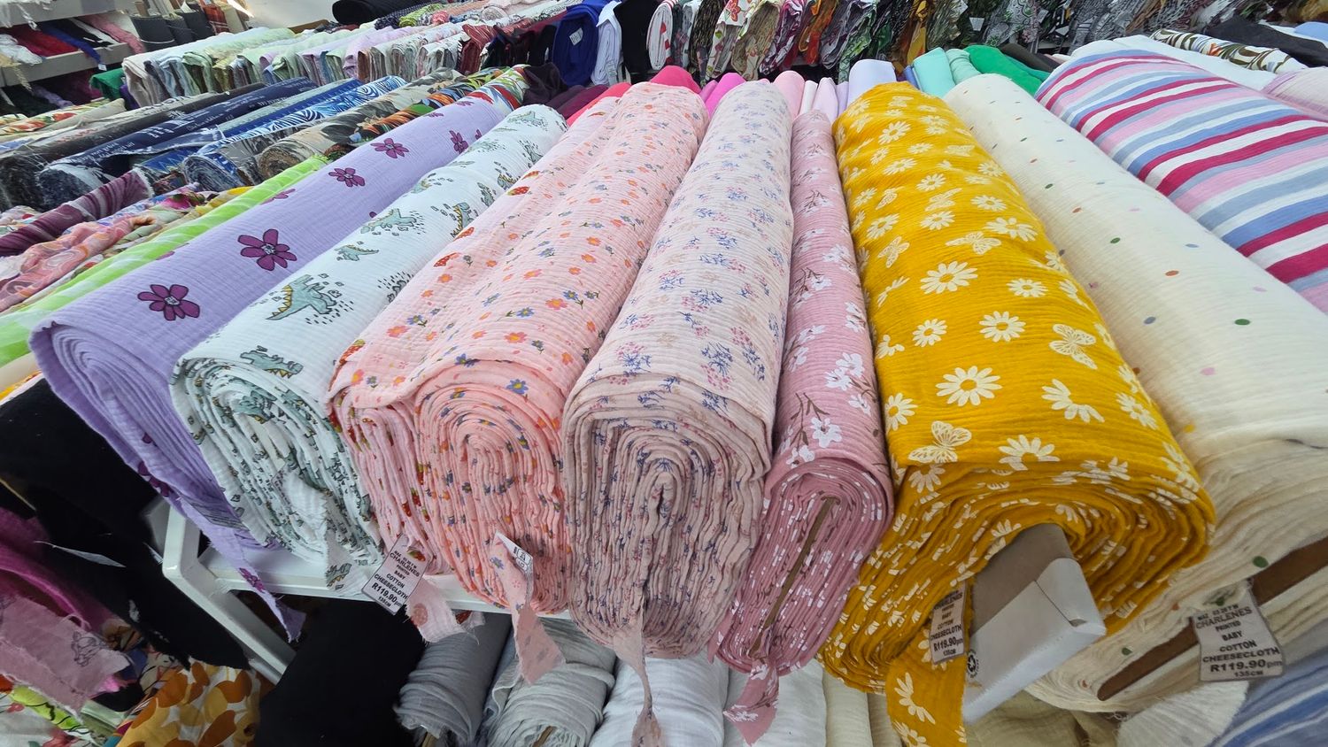 Colorful rolls of patterned fabric displayed on a market stall, featuring floral and printed textiles in pink, yellow, and purple.