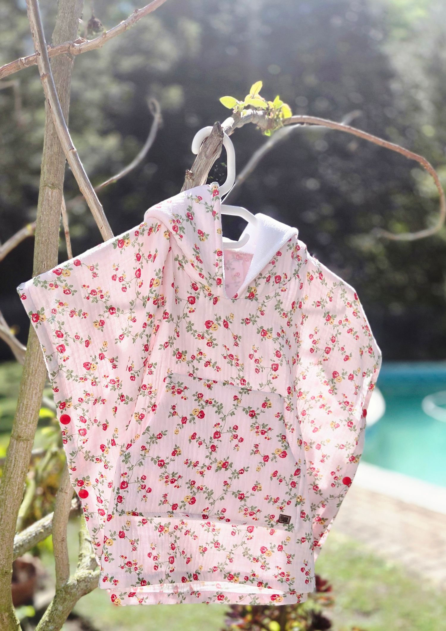 Pink floral hooded jacket hanging on a branch outdoors near a pool, featuring red rose print on white fabric.