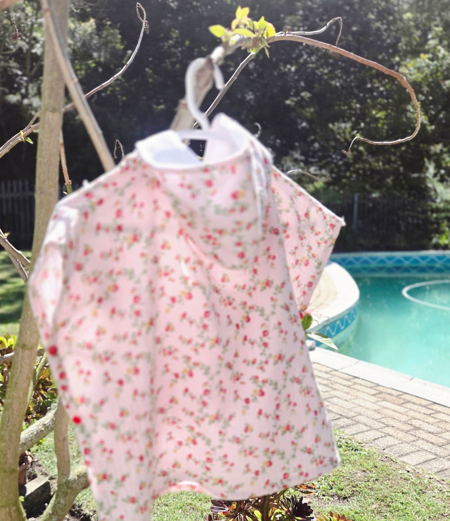 Pink floral baby dress hanging on a branch hanger outdoors near a swimming pool on a sunny day.