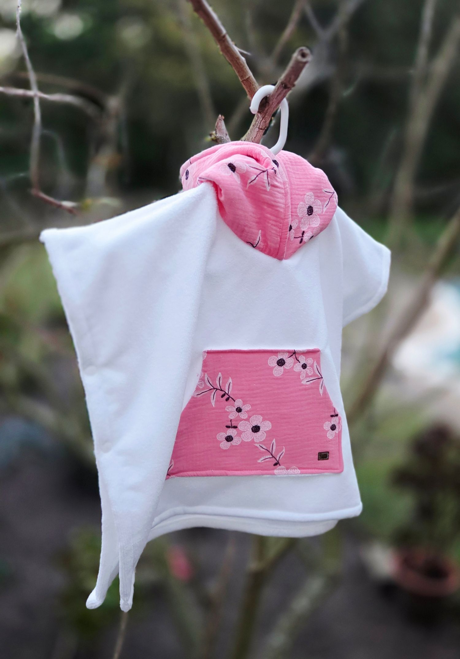 White and pink floral baby hoodie poncho hanging on a tree branch outdoors.
