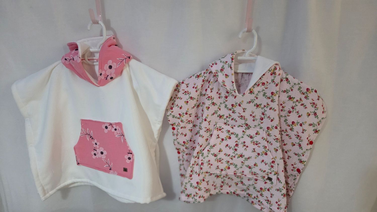 Two handmade girls pink floral hooded ponchos hanging on hangers, one white with applique, one pink ditsy print.