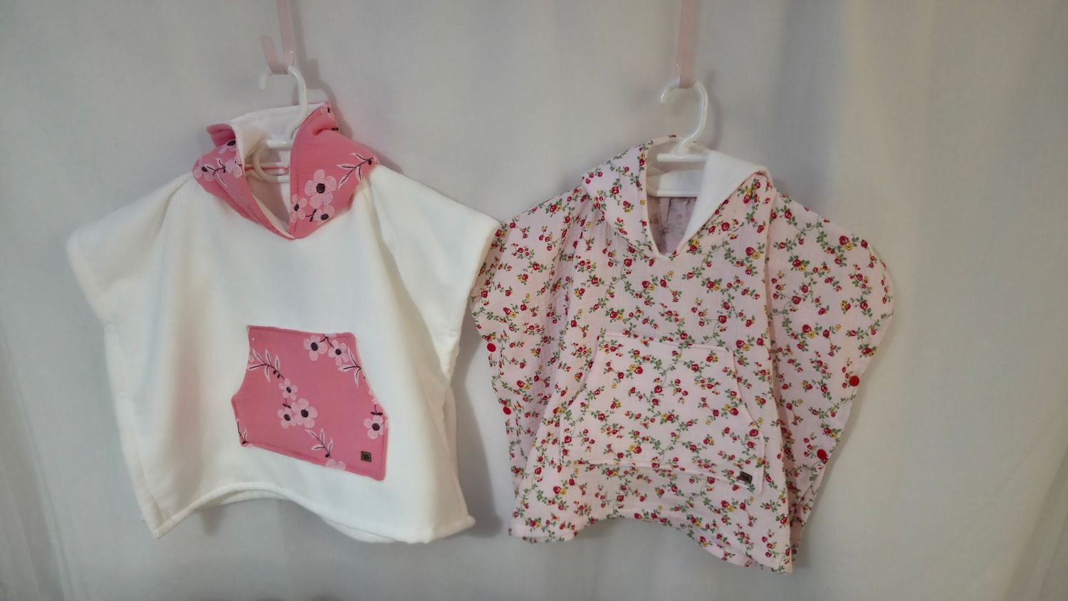 Two handmade girls poncho tops, one white with pink floral pocket and one pink floral print, hanging on display.