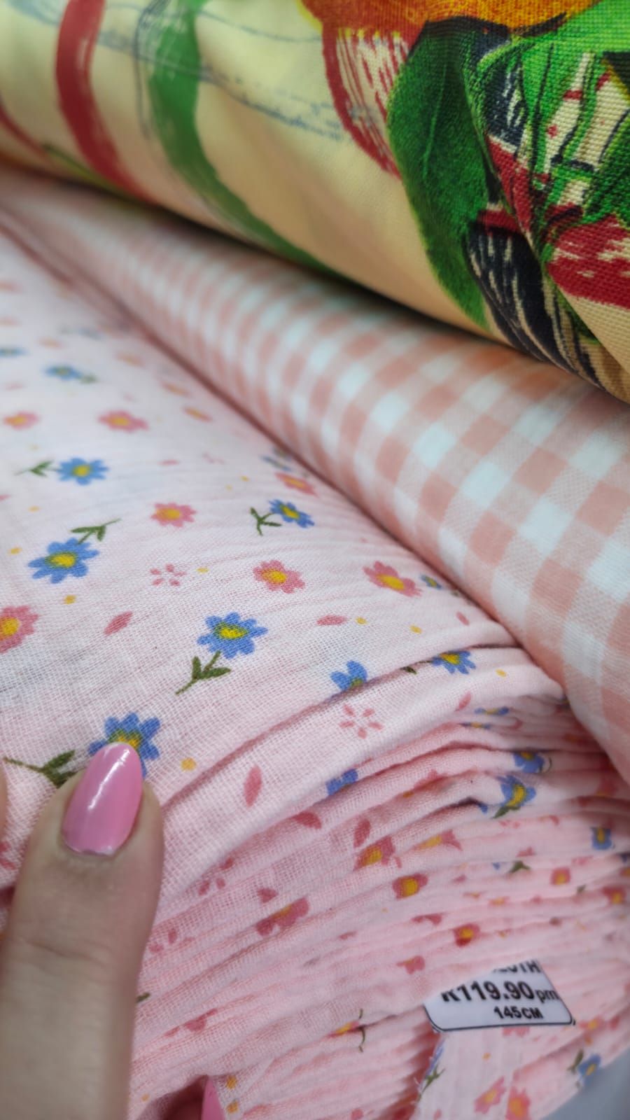 Pink floral and gingham fabric rolls displayed together, with a hand showing pink nails touching the delicate flower-print cloth.