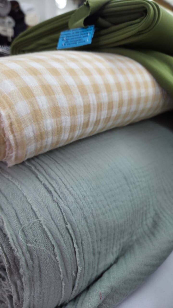 Three rolled fabric bolts stacked together featuring olive green, yellow gingham check, and sage green muslin textiles.