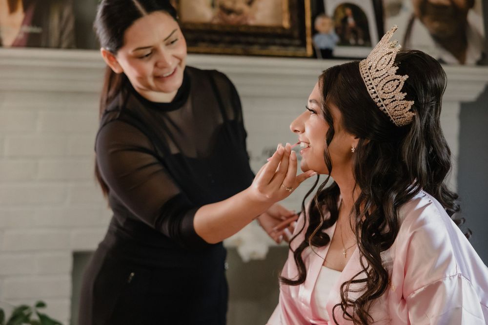 Makeup artist applying finishing touches to a quinceañera girl wearing a crown and pink robe.