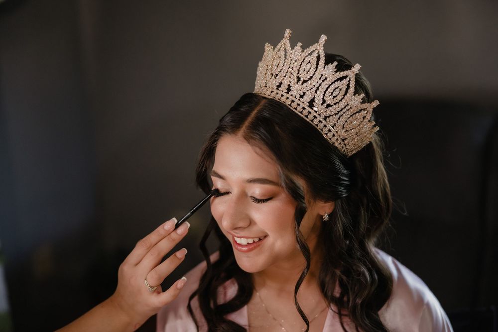 Young woman wearing gold crown gets makeup applied at her quinceañera celebration.