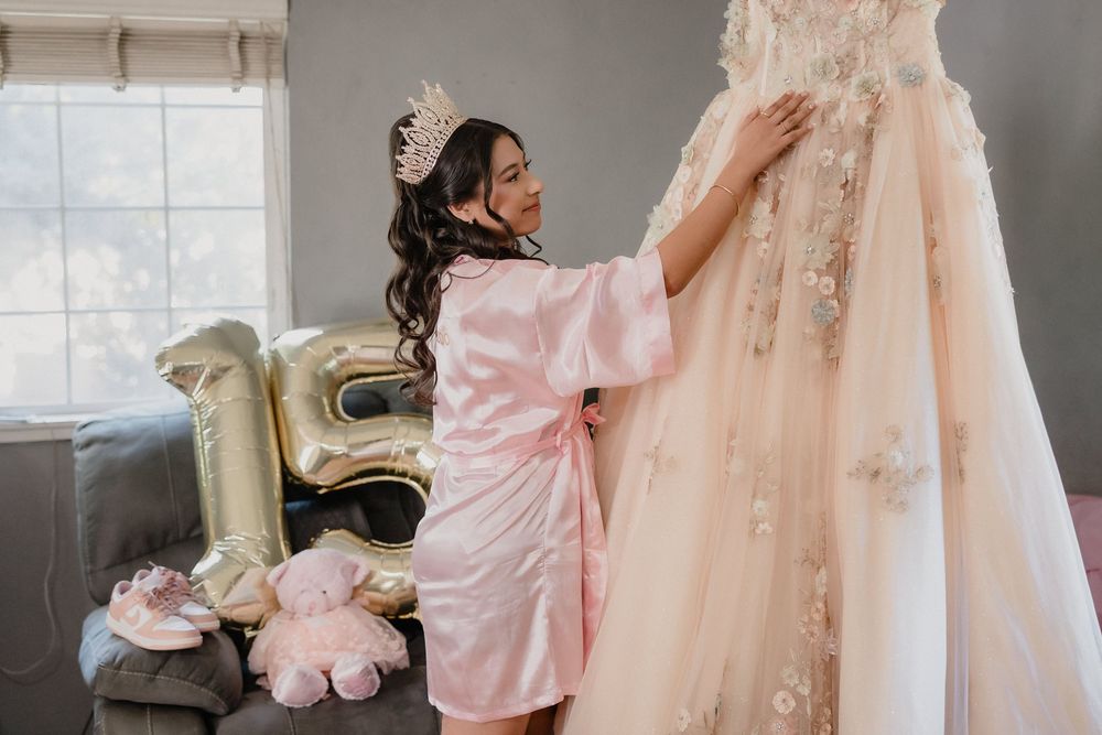 Quinceañera girl in pink robe admiring her floral ball gown hanging near gold number balloons.