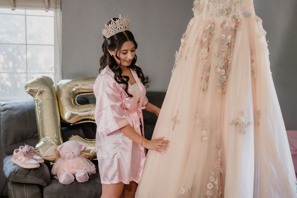 Bride in pink satin robe and tiara admires her floral ball gown quinceañera dress in a decorated room.