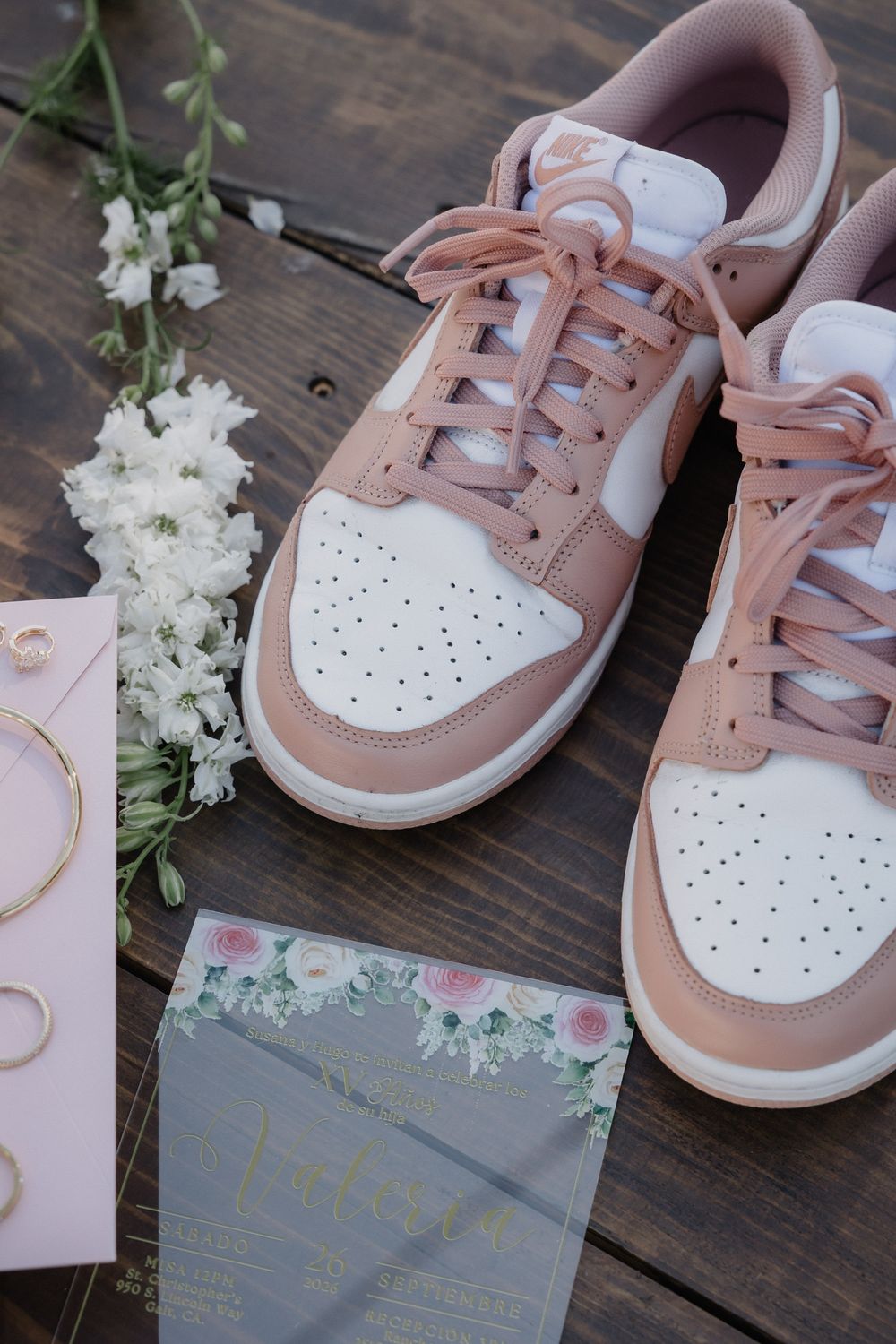 Pink Nike Dunk Low sneakers displayed with floral wedding stationery, gold jewelry, and white flowers on rustic wood.