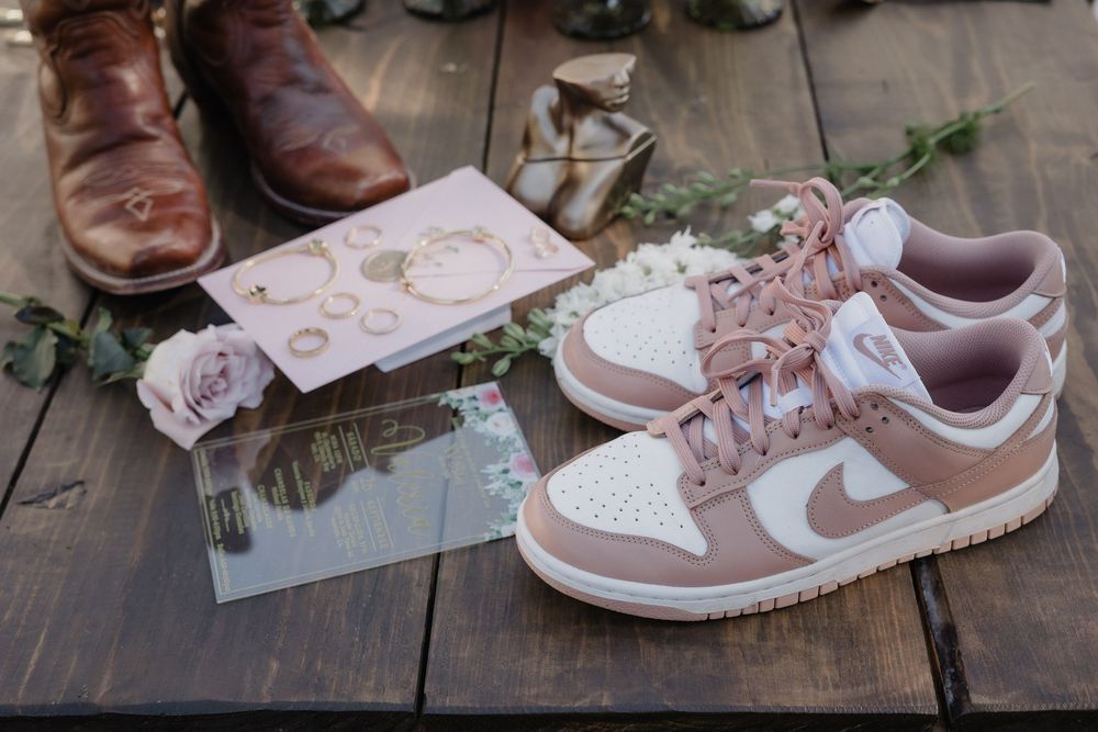 Pink Nike Dunk Low sneakers displayed with wedding rings, floral accents, and brown leather dress shoes on rustic wood.