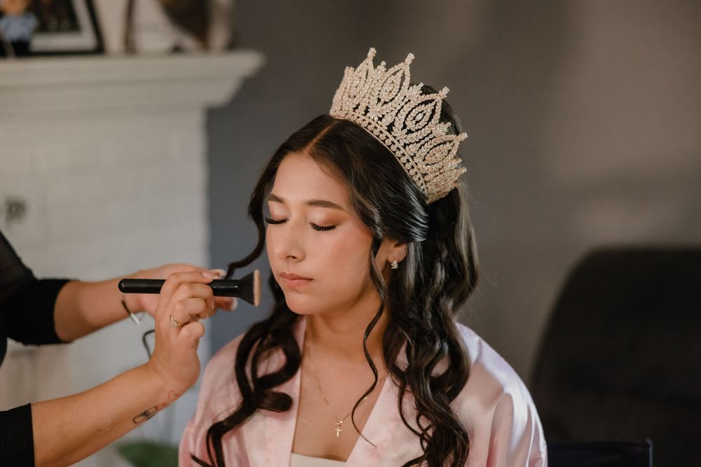 Young woman wearing a gold crown gets makeup applied during quinceañera preparations.