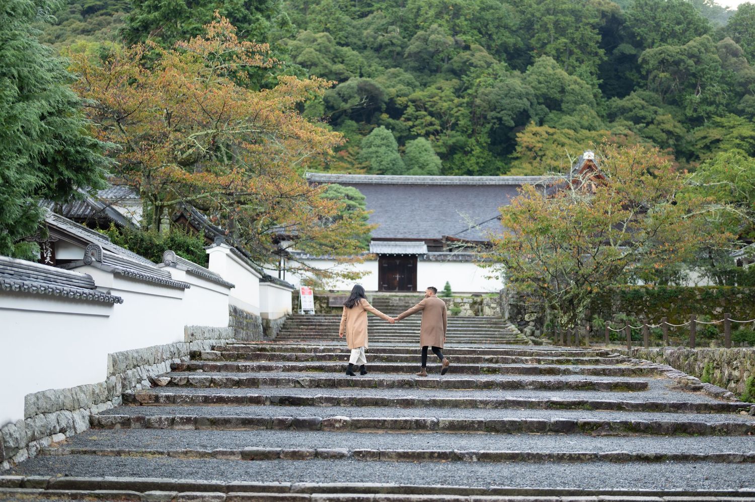 Two visitors walk up stone steps toward a traditional Japanese temple gate surrounded by lush green trees.