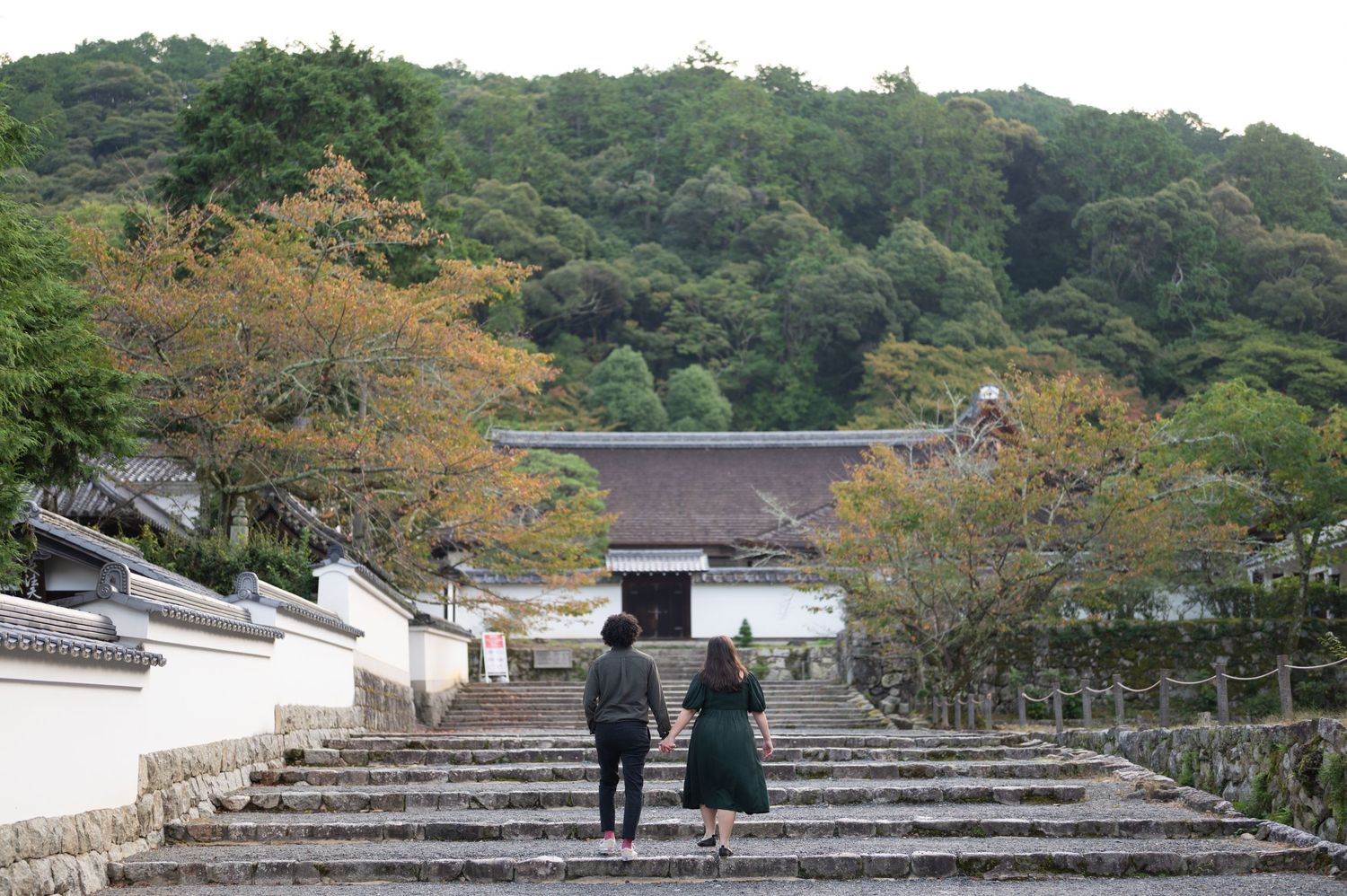 Two people holding hands walk up stone steps toward a traditional Japanese temple surrounded by lush green trees.
