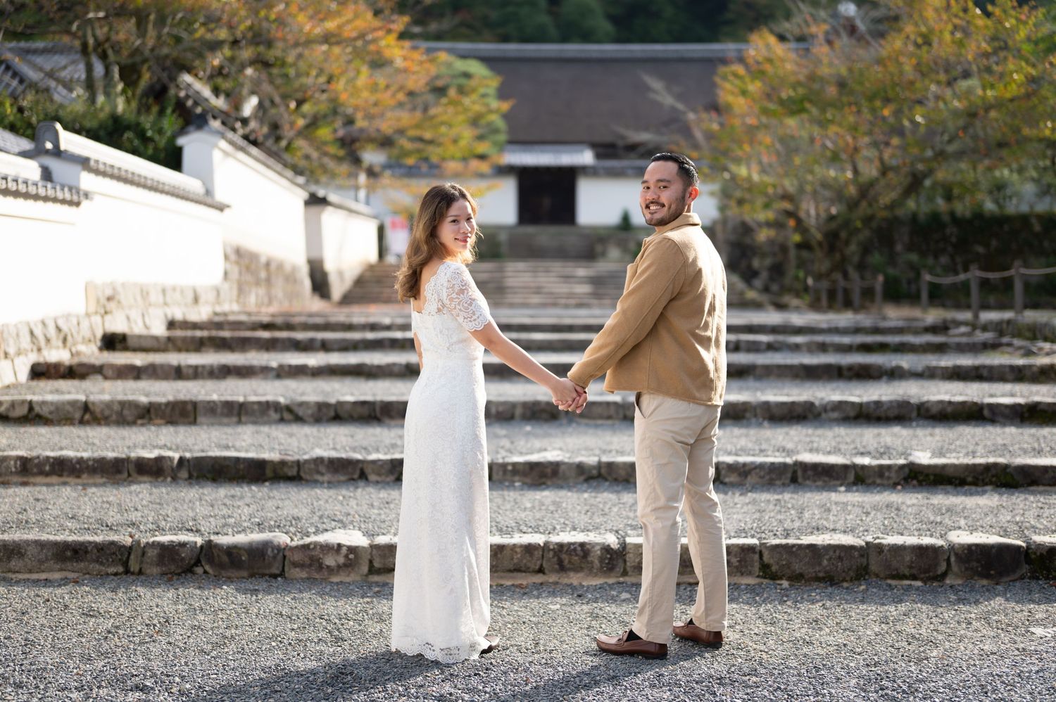 Couple in wedding attire holding hands on stone steps at a Japanese temple, smiling at camera.