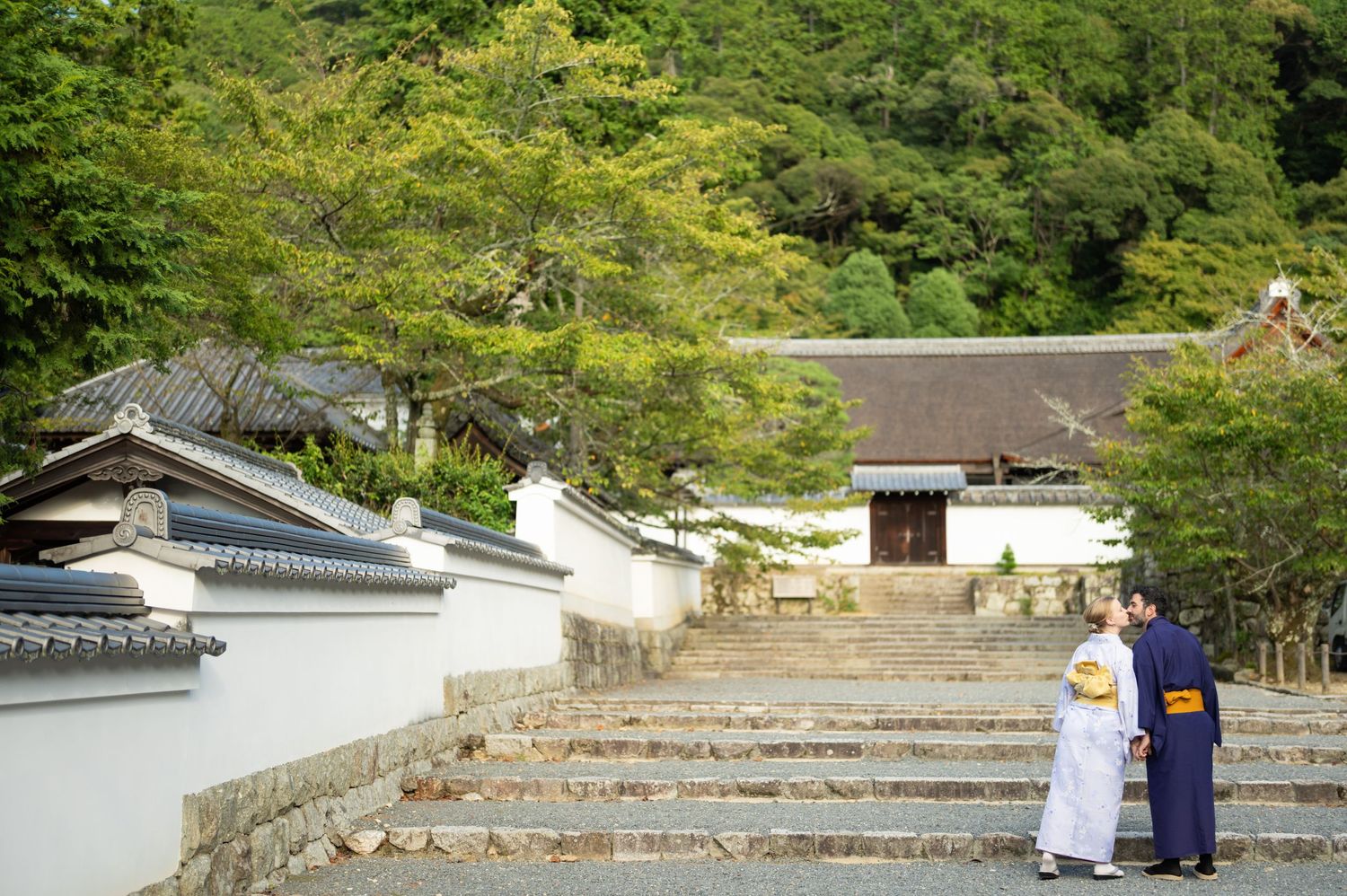 Couple in traditional Japanese kimono standing on stone steps near a historic temple surrounded by lush green trees.