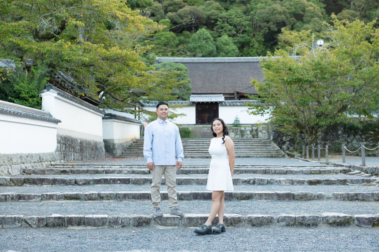 Couple standing on stone steps before a traditional Japanese temple gate surrounded by lush green trees.