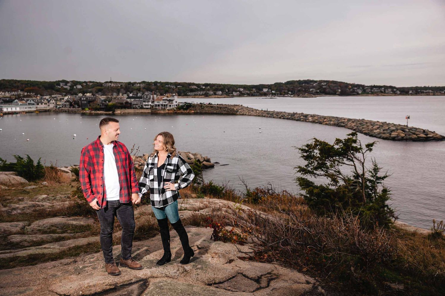 Two people standing on a rocky overlook, gazing toward the waterfront town below them. Engagement at Rockport Harbor, Rockport, MA