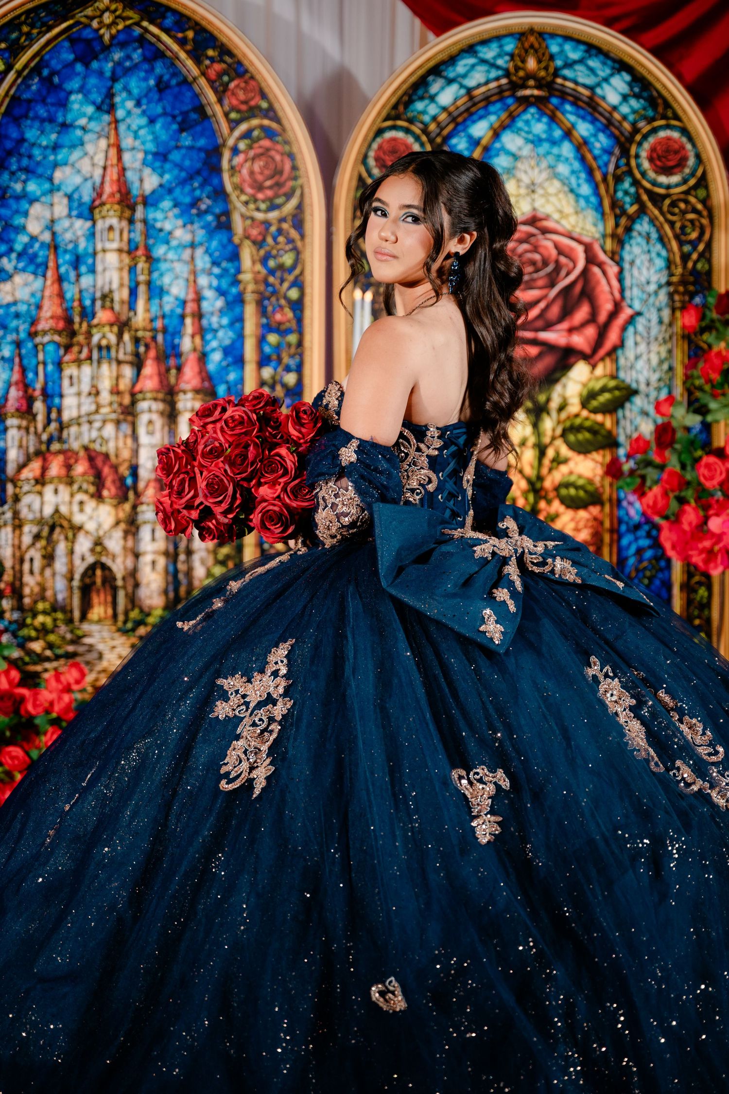 Young woman in a navy blue quinceañera ball gown with gold appliqués holding red roses before castle stained glass windows.