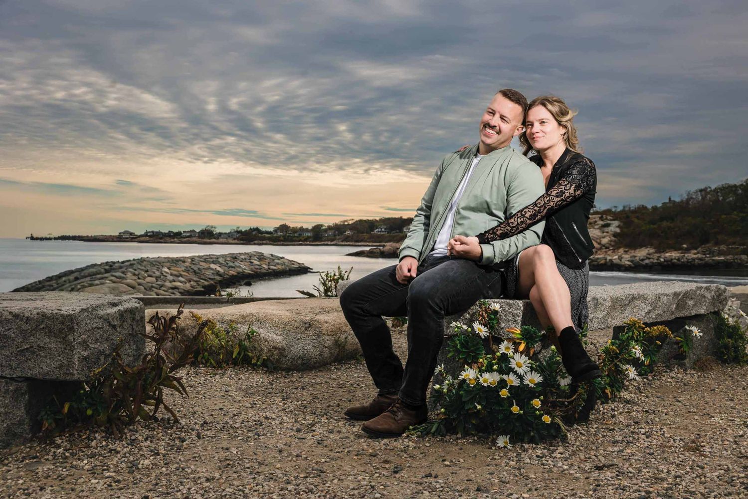 Couple sitting together on a stone wall overlooking the Atlantic smiling at the camera. Engagement at Rockport Harbor, Rockport, MA