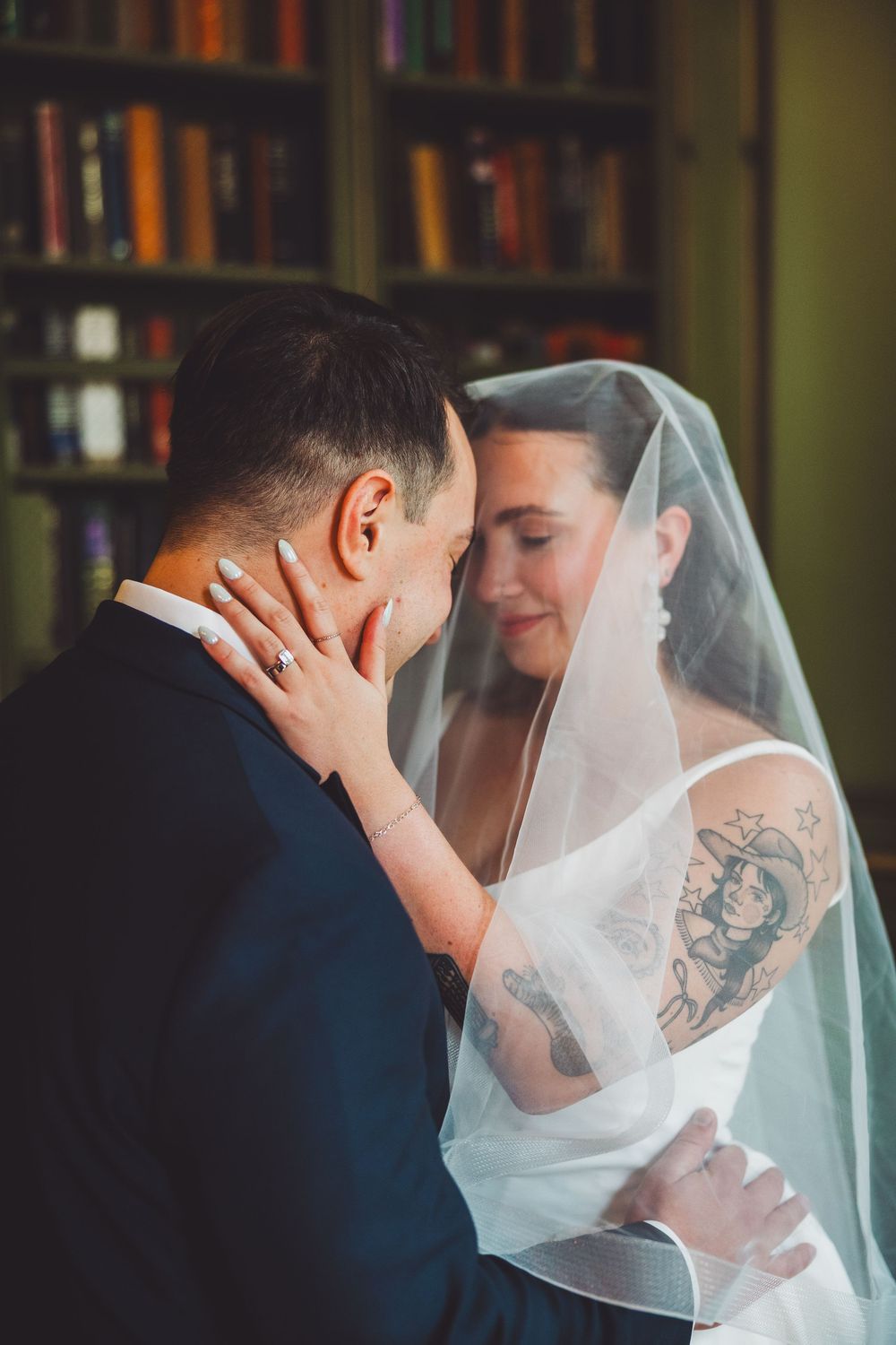Bride with tattoos and veil tenderly touches grooms face during intimate wedding moment in front of bookshelf at the meridian house