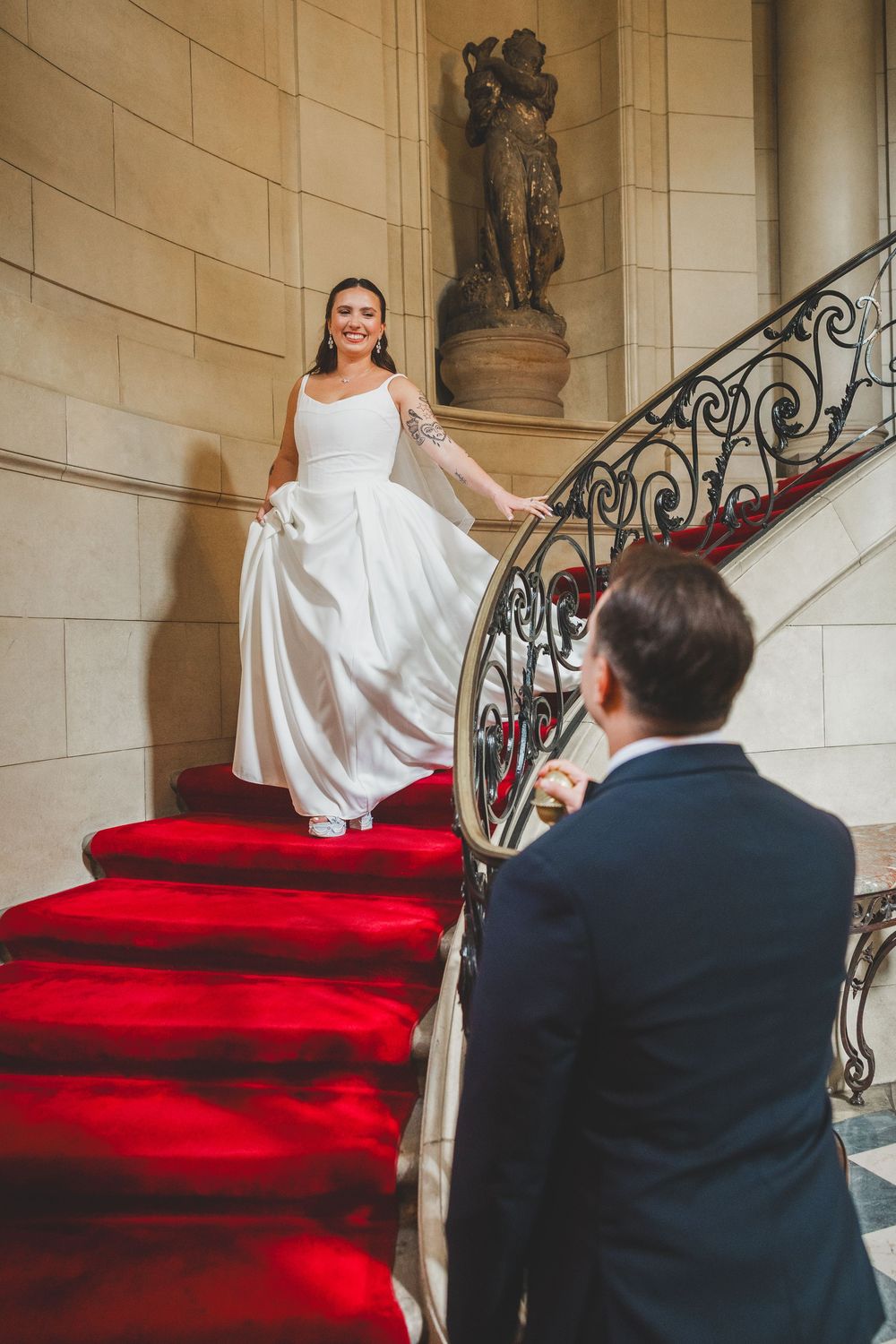 Bride in white gown descends red-carpeted staircase toward groom in navy suit, elegant venue with ornate iron railing.
