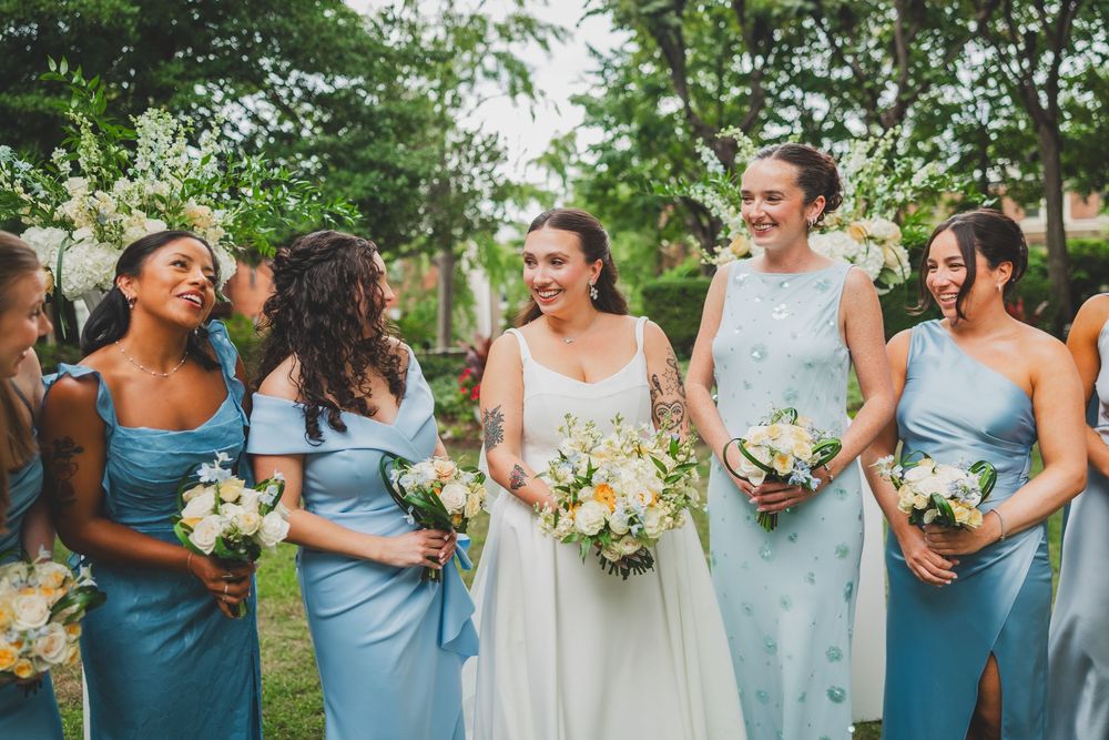 Bride in white gown walks with smiling bridesmaids in blue dresses holding bouquets outdoors at a wedding.