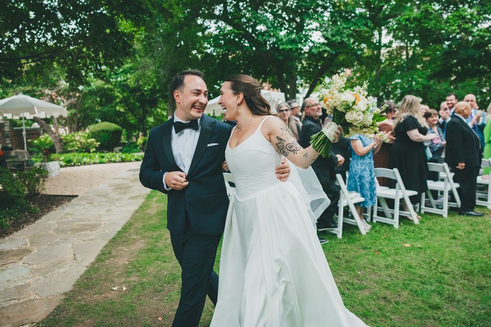 Newlywed couple smiling and walking down outdoor wedding aisle, bride in white gown holding bouquet, groom in navy tuxedo.