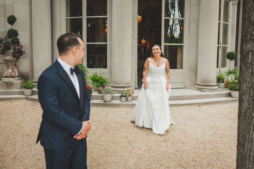 Bride in white gown approaches groom in navy tuxedo during first look at elegant outdoor venue with stone columns.