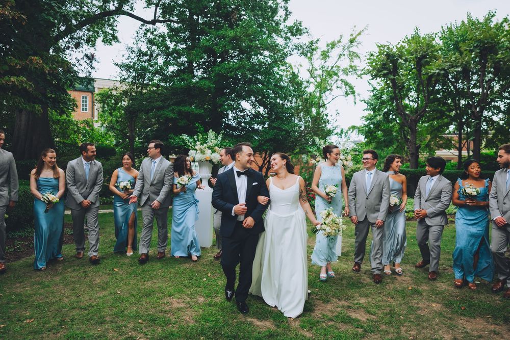 Bride and groom walk together outdoors at wedding ceremony surrounded by bridal party in blue and gray attire.