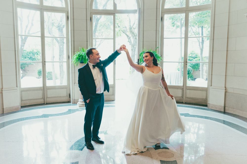 Bride and groom dancing together in elegant wedding attire inside a bright, sunlit hall with tall windows.