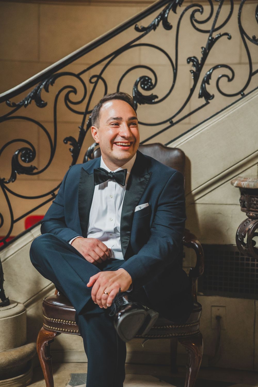 A smiling groom in a navy tuxedo with bow tie sits in a leather chair beside an ornate staircase railing.