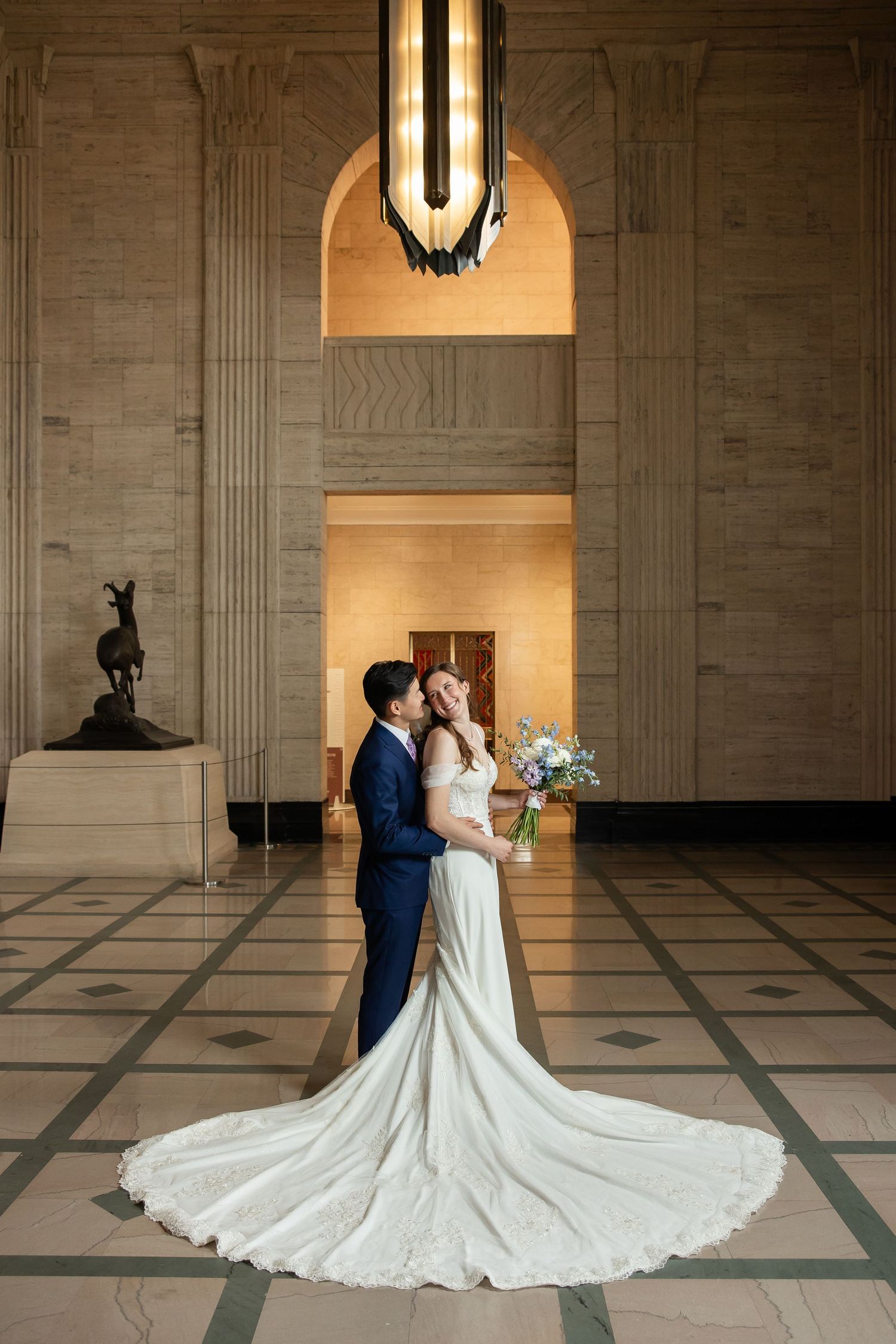Bride and groom embrace in elegant wedding venue hall with grand arched ceiling, chandelier, and flowing white bridal gown train.