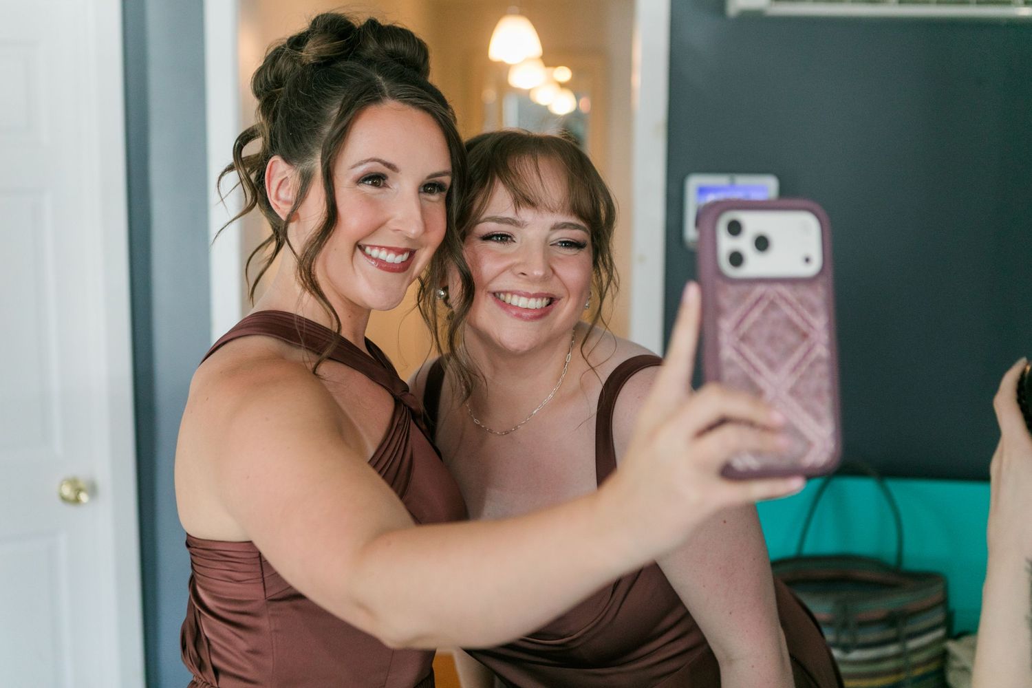 Two bridesmaids in brown satin dresses smile while taking a selfie together before a wedding ceremony.