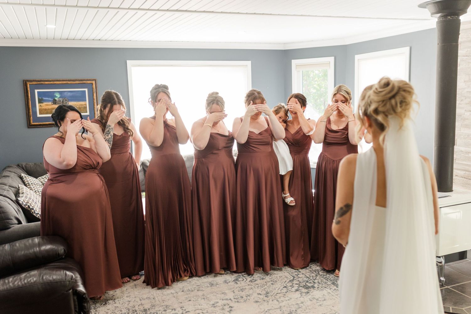 Bridesmaids in rust-colored gowns react emotionally to bride's first look reveal indoors.