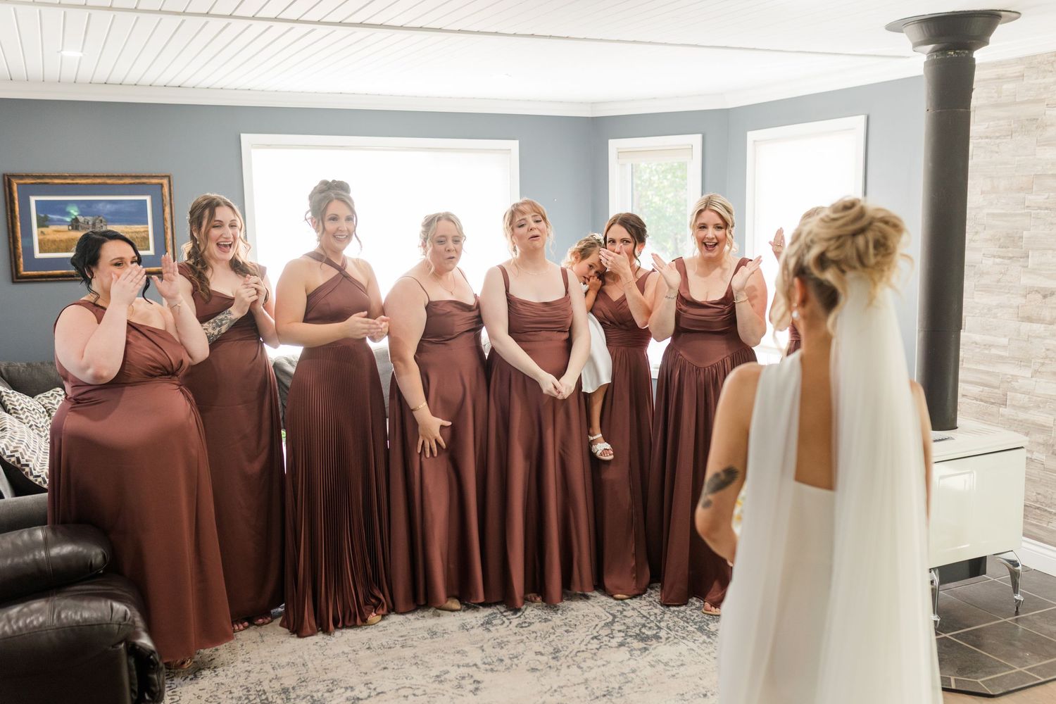 Bride reveals wedding dress to bridesmaids in rust-colored gowns, capturing emotional first look reactions indoors.
