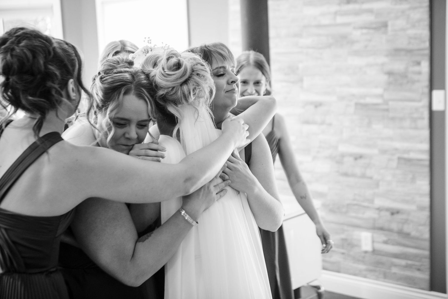 Bride and bridesmaids share an emotional group hug in a bright venue during wedding preparations.