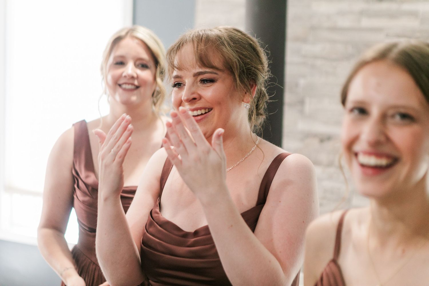 Three bridesmaids in brown dresses clapping and smiling joyfully at a wedding celebration.