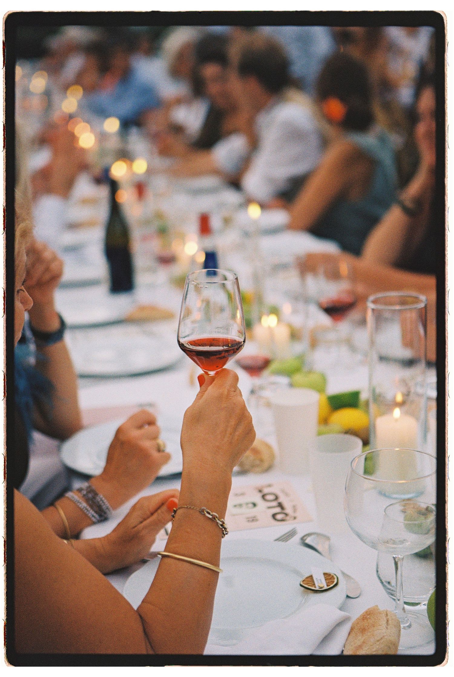 Hand holding a glass of red wine at an elegant outdoor dinner party with candles and guests in background.