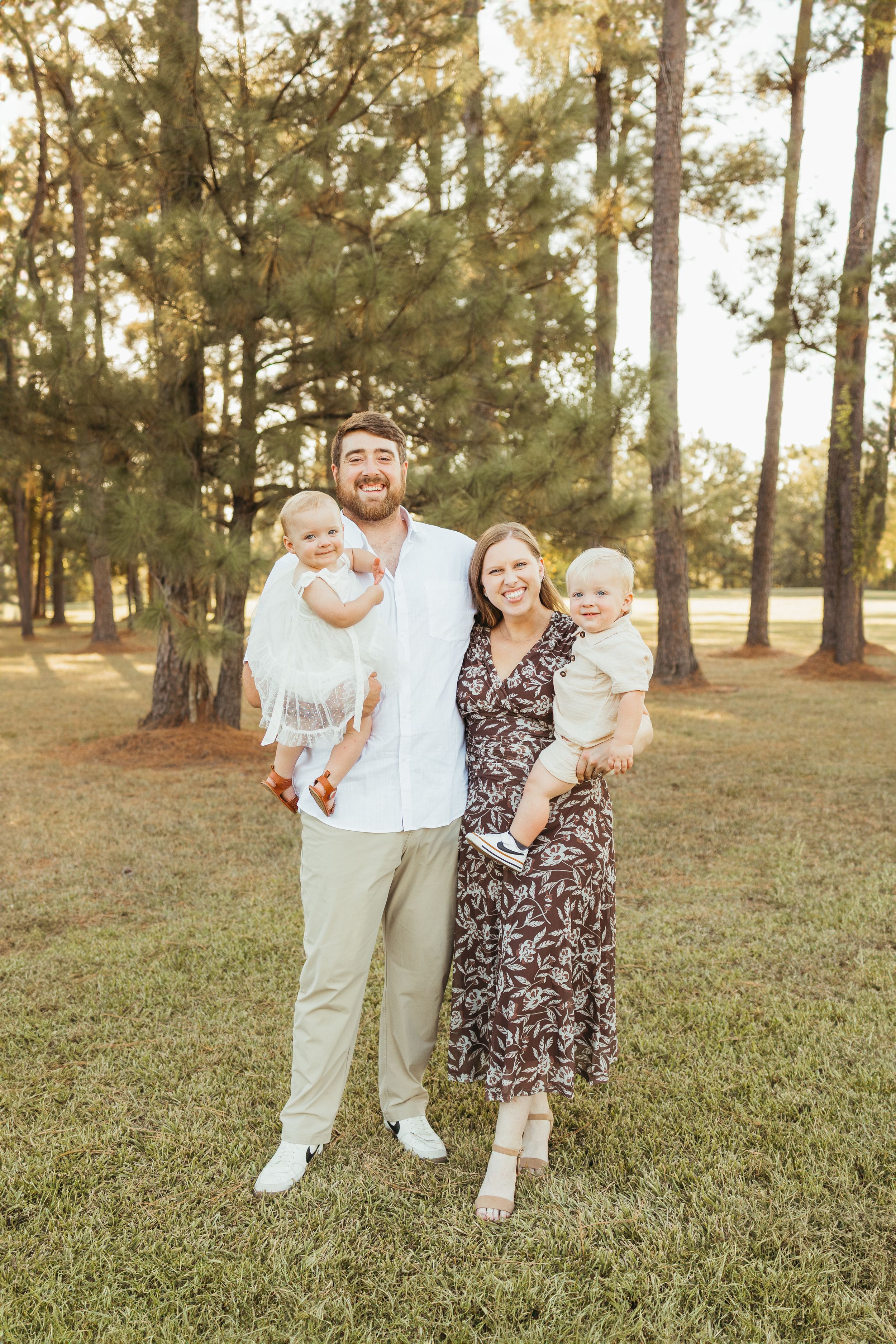 Young family with two babies posing outdoors in a sunny pine tree park during golden hour In Friendswood Texas
