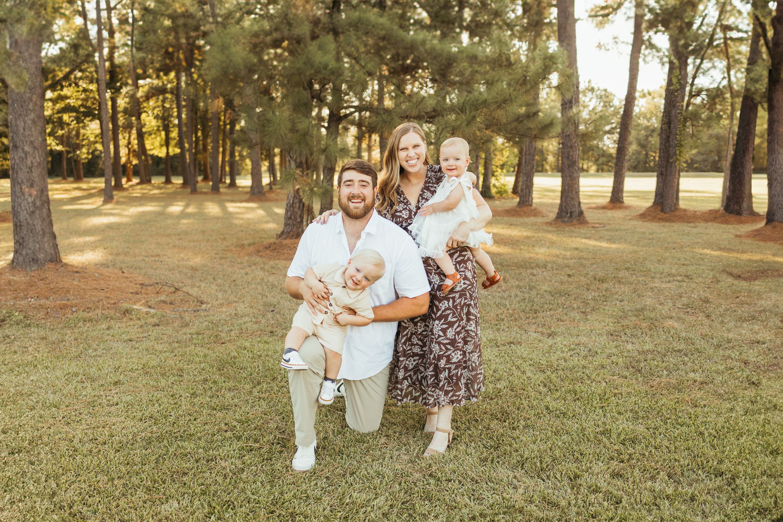 Family of three posing outdoors in a pine tree park, parents holding toddler and puppy on green grass In Friendswood Texas