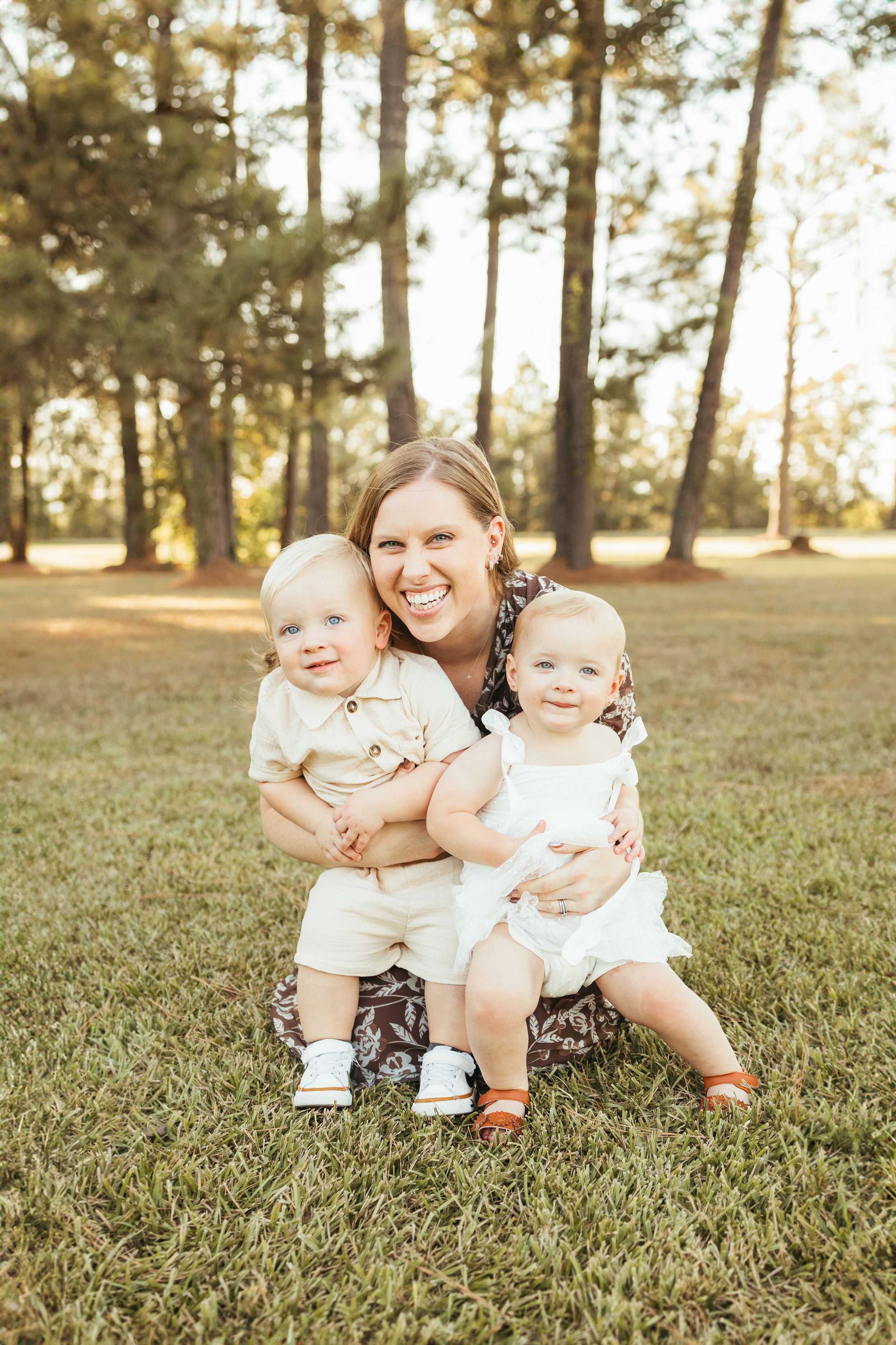 A smiling woman sits on green grass hugging two happy toddlers outdoors with tall pine trees in the background In Friendswood Texas