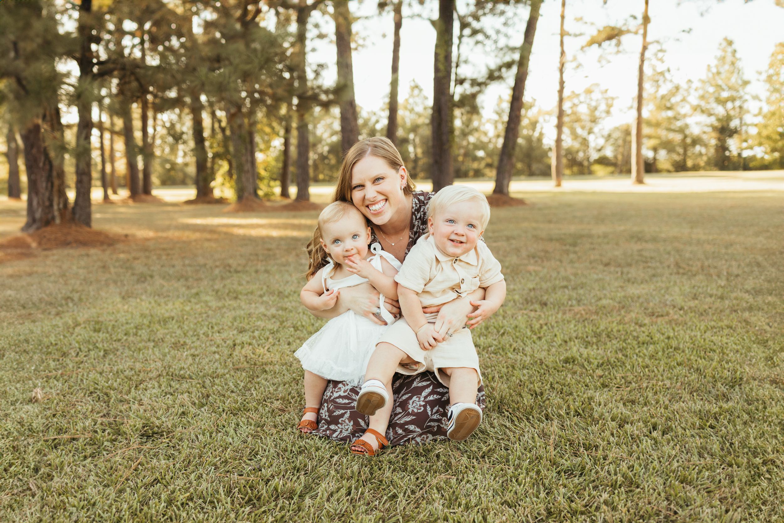 A smiling woman sits on grass in a sunny park, hugging two young toddlers in white outfits, surrounded by tall trees In Friendswood Texas