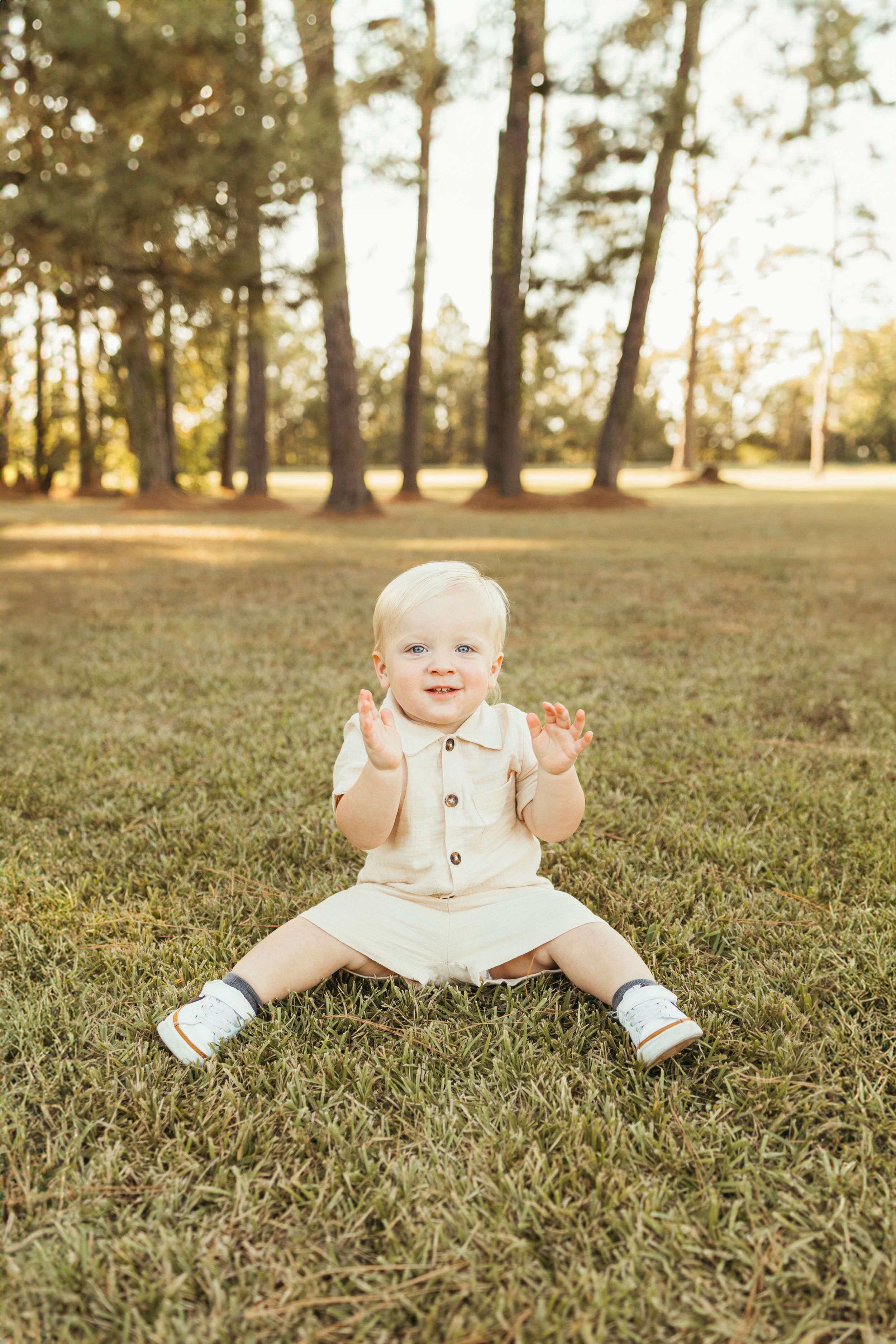 An adorable blonde toddler boy in a cream romper claps hands while sitting on green grass in a park In Friendswood Texas
