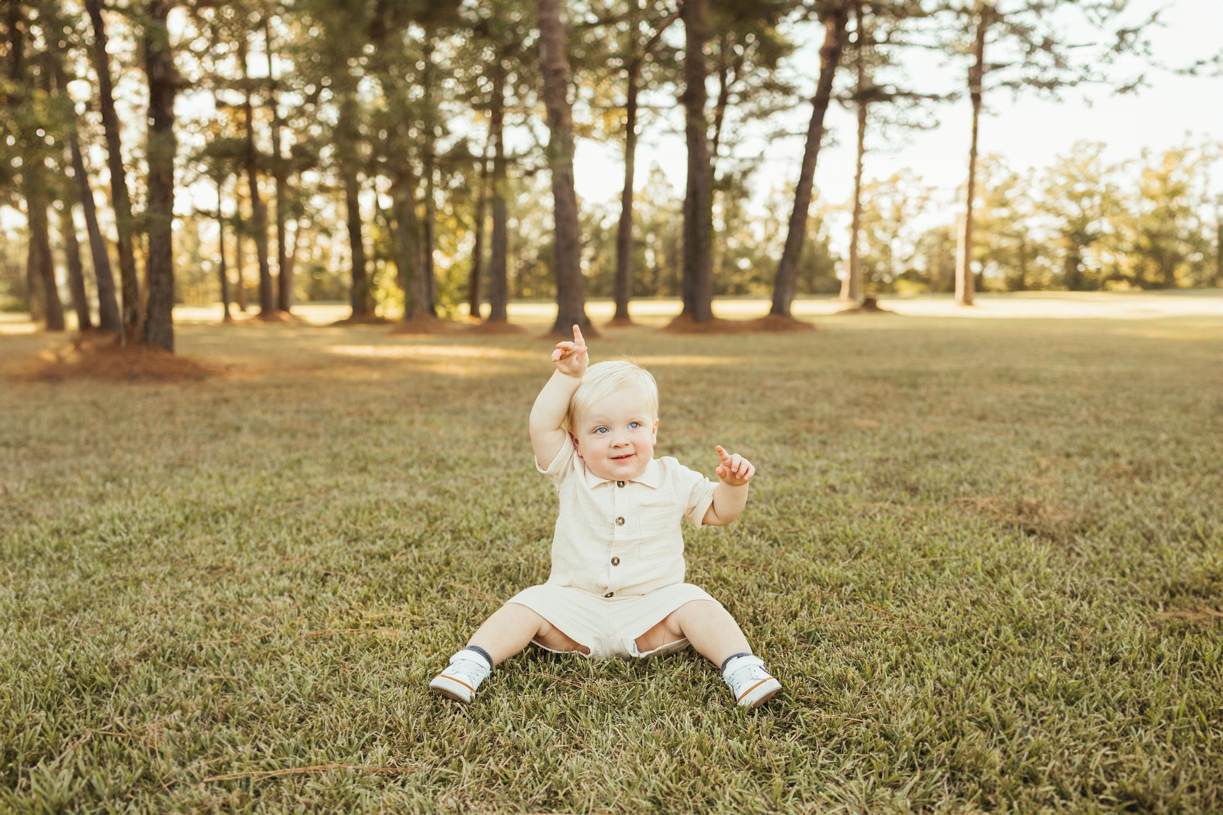 Adorable baby in cream outfit sitting on grass in a sunny park, raising one arm joyfully In Friendswood Texas
